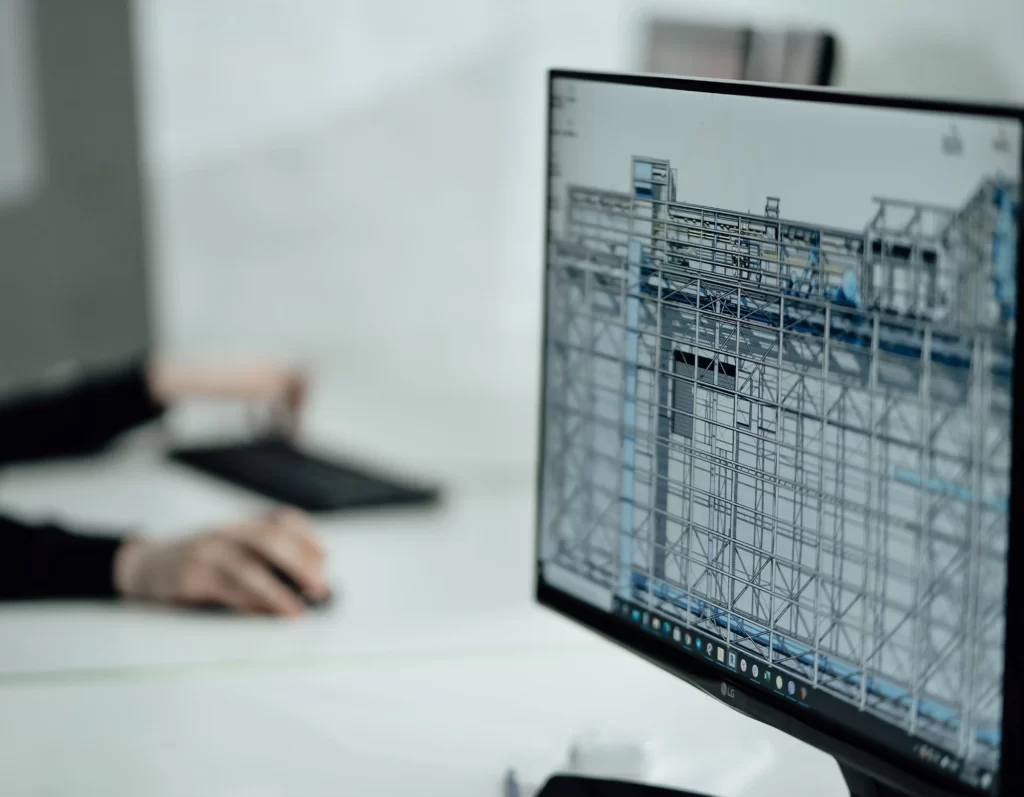 Monitor en primer plano con im├ígen de autocad. De fondo dos manos trabajan con teclado y mouse.
